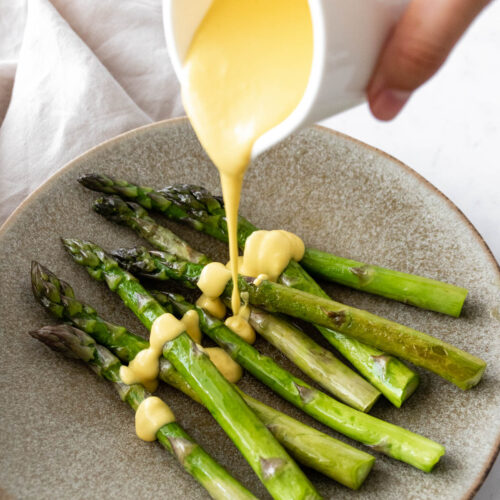 A plate with asparagus and a small jug pouring Hollandaise sauce over the asparagus