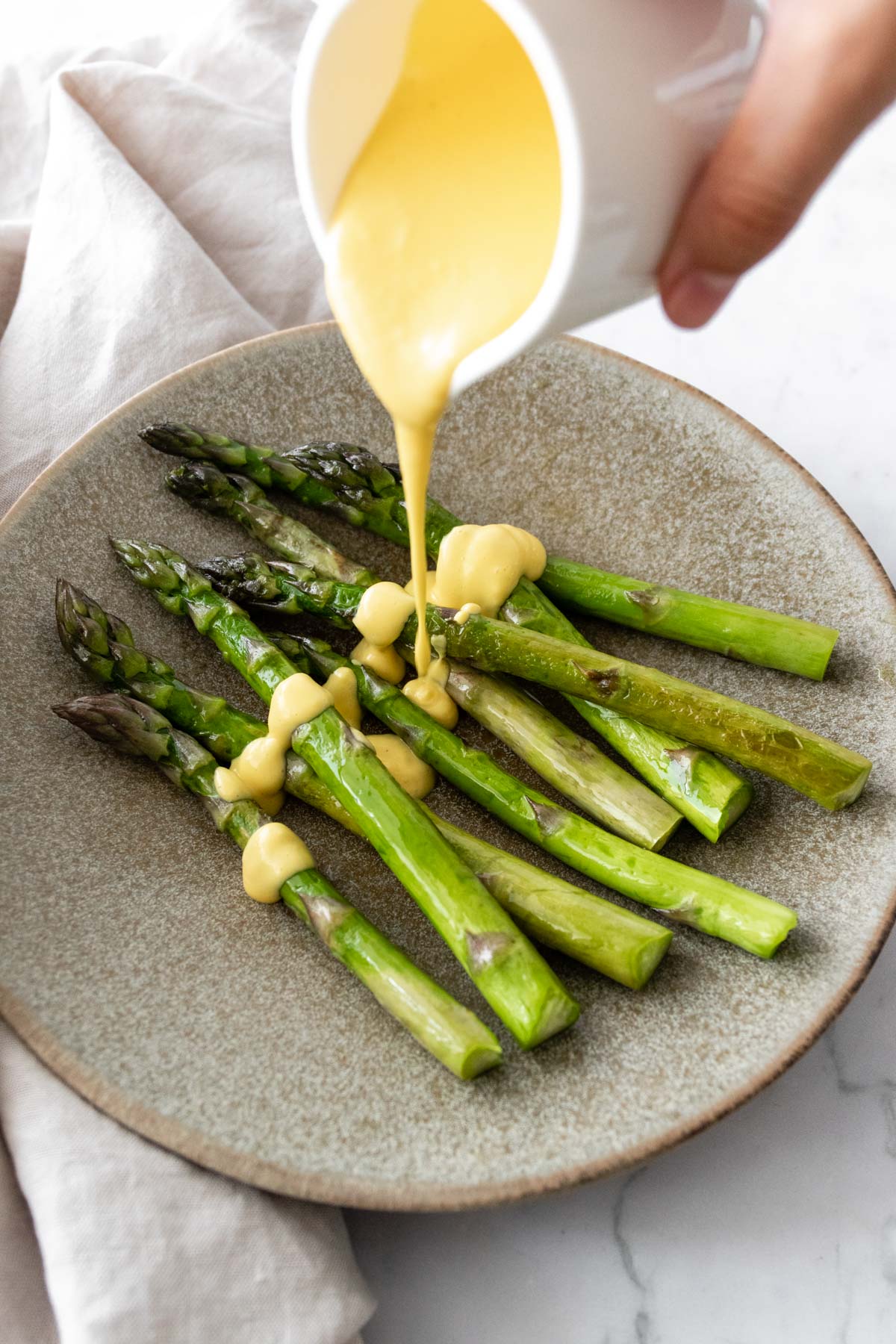 a plate with asparagus and a hand pouring Hollandaise sauce over the asparagus