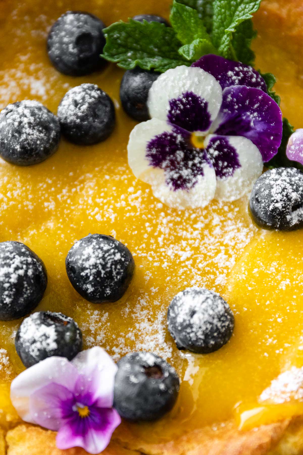 close up to a dutch baby pancake with lemon curd, blueberries, mint, wildflowers, and powdered sugar